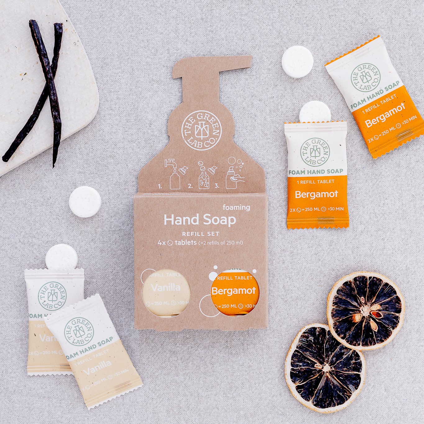 The Green Lab Co Foaming Hand Soap Refill Set (4 tablets), Bergamot and Vanilla, with loose tablets, dried citrus slices, and vanilla pods on a light fabric.